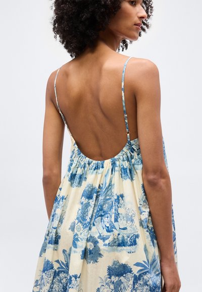 Γυναίκα με σγουρά μαλλιά, φορώντας φόρεμα μπλε και κρεμ με λουλούδια, χωρίς πλάτη, με λεπτές ρυθμιζόμενες τιράντες, σε προφίλ μπροστά σε απλό φόντο.