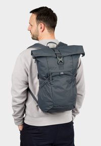 Grauer Rolltop-Rucksack mit verstellbaren Riemen, Schnallenverschluss und strukturiertem Stoff. Verfügt über eine Seitenasche und ein minimalistisches Design.