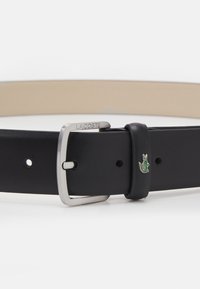 Zwarte leren riem met een zilveren metalen gesp en een groen krokodillenlogo. Heeft een gladde textuur met meerdere verstelgaten.