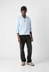 Suéter de manga larga azul claro con cremallera, combinado con jeans negros y zapatillas beige. El conjunto incluye una gorra negra.