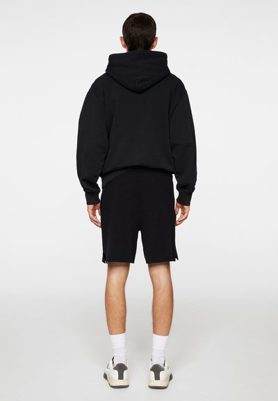 Svart hettegenser med løs passform, parret med svarte shorts. Begge plaggene har myk stoffkvalitet og minimal detaljering. Hvite sokker og sneakers.