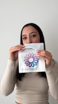Joven mujer con cabello largo y oscuro sostiene y muestra un paquete de mascarilla facial blanca con un diseño espiral colorido sobre un fondo liso.
