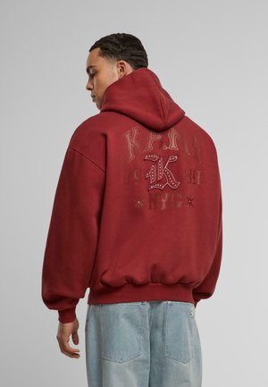Maroon oversized hoodie with a front pocket, featuring embossed text and graphics in a contrasting shade. Soft fabric with ribbed cuffs.
