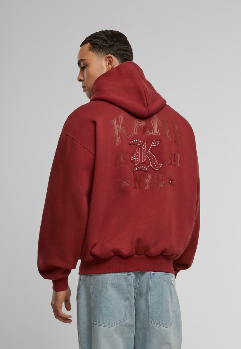Maroon oversized hoodie with a front pocket, featuring embossed text and graphics in a contrasting shade. Soft fabric with ribbed cuffs.