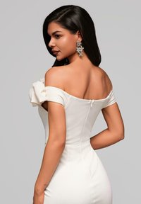 Robe blanche à épaules dénudées à coupe ajustée, avec un accent floral sur l'épaule et une fermeture éclair au dos. Boucles d'oreilles métalliques visibles.