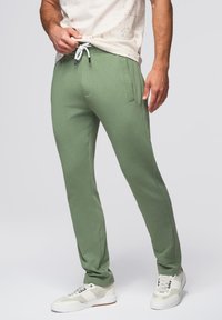 Groene sweatpants met een ontspannen pasvorm, een trekkoord in de taille, zijzakken en een zachte textuur. Gecombineerd met witte en grijze sportschoenen.