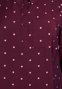 Bordeauxfärgad skjorta med små rosa prickar, har en bröstficka, krage och textilstruktur. Knapparna matchar skjortans färg.