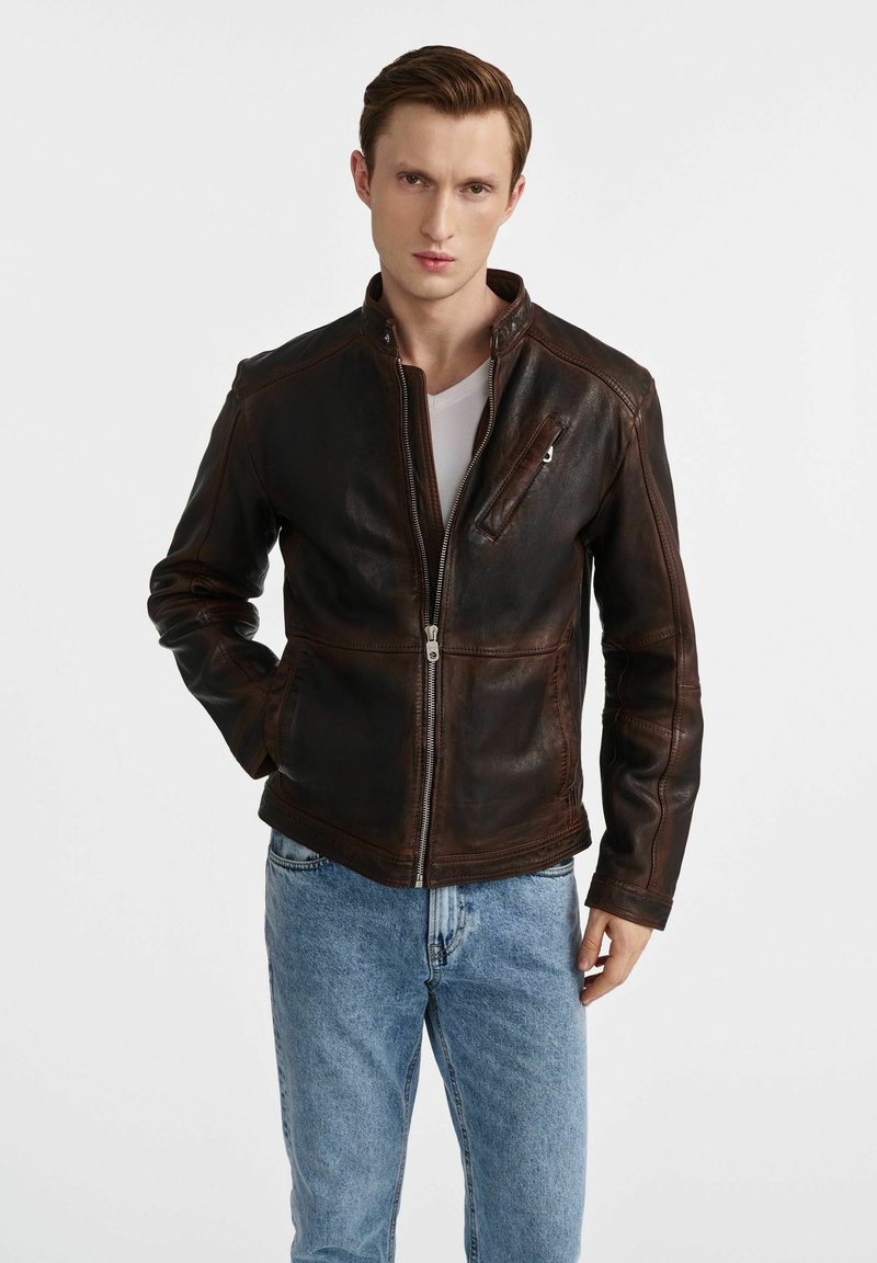 Brązowa skórzana kurtka z zamkiem z przodu, wysokim kołnierzem i bocznymi kieszeniami. Posiada małą kieszeń na zamek na klatce piersiowej oraz fakturowane wykończenie.