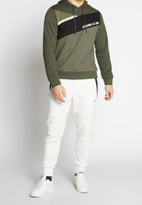 Felpa con cappuccio in verde oliva e nero con una striscia bianca, caratterizzata da un logo. Abbinata a pantaloni joggers grigi chiari e sneakers bianche.