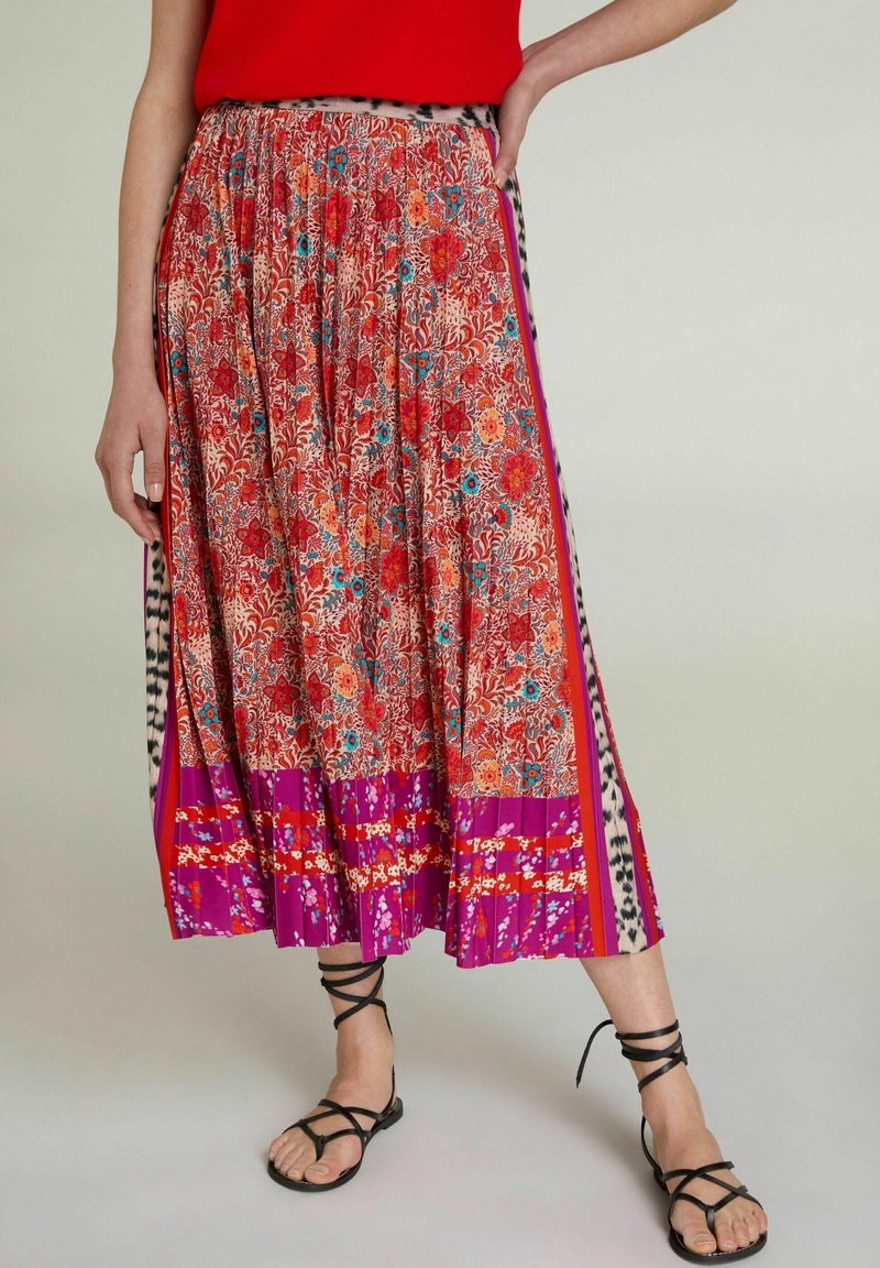 Femme portant un haut rouge et une jupe midi plissée à fleurs colorées avec des sandales noires lacées, debout avec une main sur la hanche.