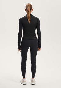 Black athletic top with a high collar, fitted design, and textured diagonal stitching paired with black leggings and light sports shoes.