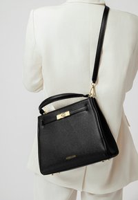 Borsa a mano nera in pelle con una forma strutturata, accessori in oro, superficie testurizzata e tracolla removibile, appoggiata su un outfit chiaro.