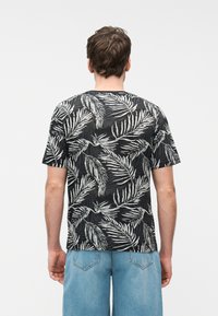 Άνδρας με την πλάτη γυρισμένη, φοράει μαύρο κοντομάνικο πουκάμισο με λευκό μοτίβο φύλλων και ανοιχτό μπλε τζιν σορτς, μπροστά σε απλό φόντο.