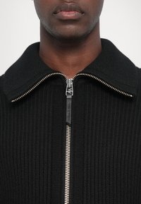 Fekete bordás kötött zipzáras dzseki magas gallérral, ezüst színű zipzár hardverrel és fekete bőr húzózsinórral, kiemelve a textúrált designt.
