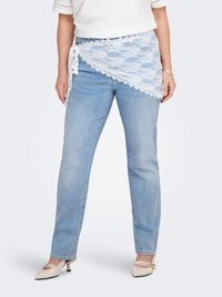 Γυναίκα που φοράει ανοιχτό μπλε τζιν και λευκό δαντελένιο ζακέτο δεμένο στη μέση, συνδυασμένο με κρεμ παπούτσια με χαμηλό τακούνι και λευκό τοπ.