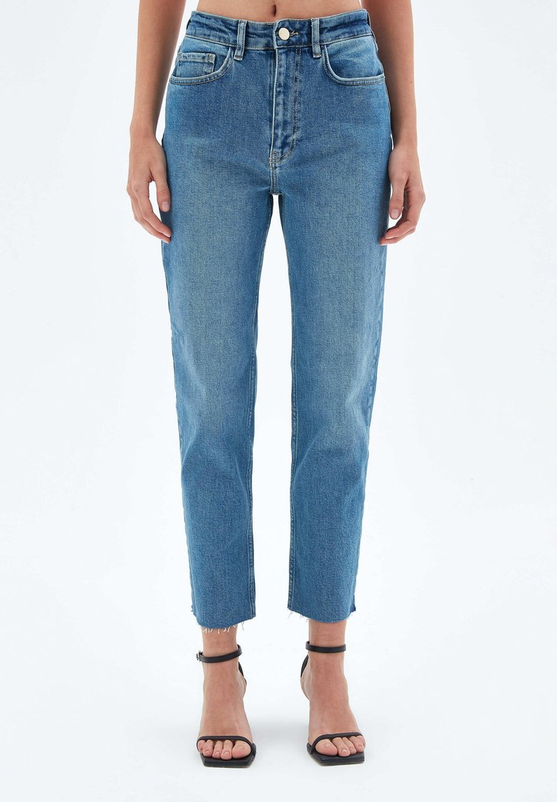 Femme portant un jean bleu taille haute, ajusté, avec des ourlets bruts et des sandales noires à bride cheville et à bout ouvert, debout devant un fond uni.