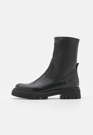 Bota de tobillo de cuero negro con punta redondeada, paneles laterales elásticos y suela de goma texturizada con ranuras profundas para mayor tracción.