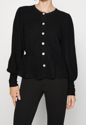 Femme portant un cardigan noir texturé avec des manches bouffantes, un col rond avec un bord argenté et des boutons ronds brillants, associé à un pantalon noir.