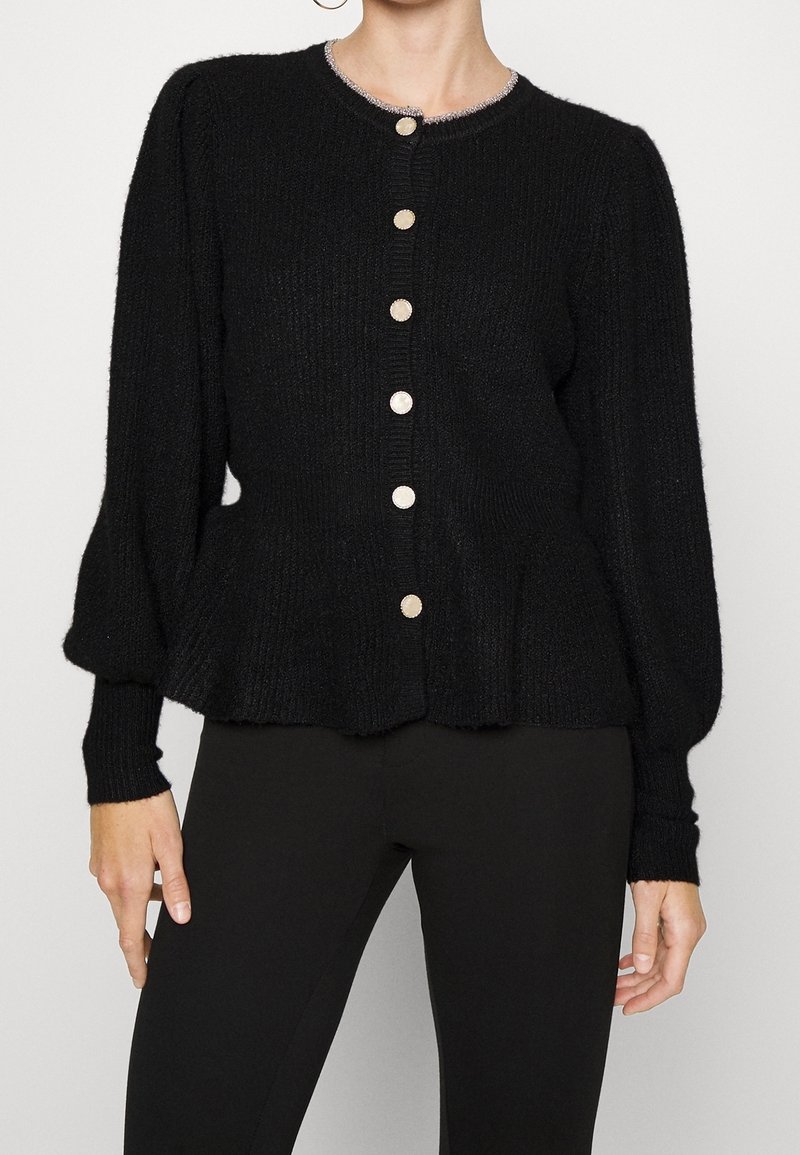 Femme portant un cardigan noir texturé avec des manches bouffantes, un col rond avec un bord argenté et des boutons ronds brillants, associé à un pantalon noir.