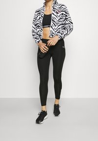 Biała i czarna kurtka z wzorem zebry z zamkiem błyskawicznym, czarny stanik sportowy, czarne legginsy i czarne buty sportowe. Butelka wody trzymana w prawej ręce.