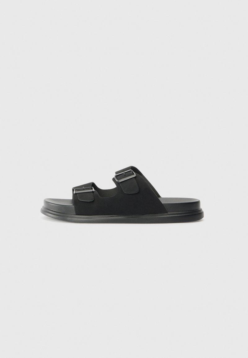 Black slide sandals feature two adjustable straps with metal buckles, a flat sole, smooth texture, and a rounded shape.