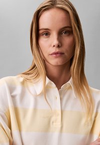 Jeune femme aux cheveux blonds longs portant un polo à rayures blanches et jaune pâle, expression neutre, sur un fond gris uni.