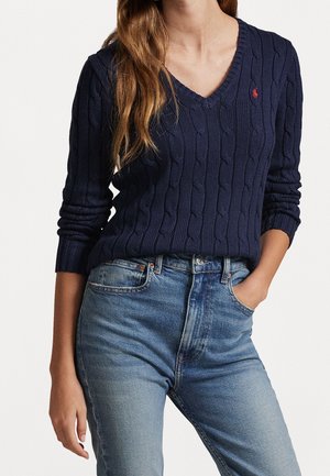 Femme portant un pull en maille torsadée bleu marine à col en V et un jean taille haute bleu clair, avec une main dans la poche, sur fond uni.
