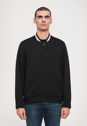 Chaqueta bomber negra con cremallera y cuello acanalado, con un discreto parche de logo. Fabricada en tejido suave, con bolsillos laterales y un ajuste relajado.