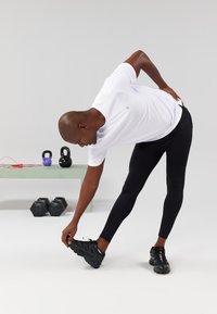 Juodos sportinės leginsai ir balti trumpomis rankovėmis marškiniai, kuriuos užsima žmogus, atliekantis tempimo pratimus. Fone matomi svoriai ir keturlenkiai ant žalios sofos.