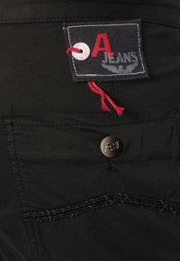 Svarta jeans med ett tygmärke med röd text "A JEANS", en vit ögla och en silverfärgad knappdetalj. Synlig sömnadsdetalj på fickorna.