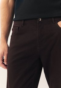 Mörkbruna byxor i mjukt tyg med smal passform, bälteshällor, knappstängning och sidofickor med kontrasterande sömmar.