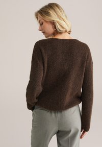Pulover călduros, maro, cu un croi lejer și gât larg, însoțit de pantaloni gri deschis, vizibili din spate.