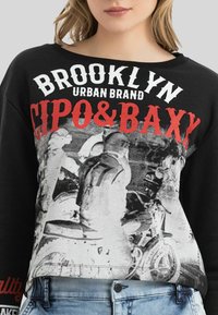 Schwarzer Sweatshirt mit oversized Passform, der weißen und roten grafischen Schriftzüge zeigt. Im Zentrum ist ein Graustufenbild mit Motorrädern zu sehen.