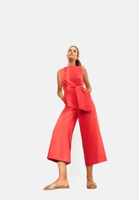 Tuta rossa con un corpetto aderente, ampie gambe a culotte e un dettaglio a fiocco frontale. Il materiale sembra liscio e resistente. Accompagnata da sandali piatti e decorati.
