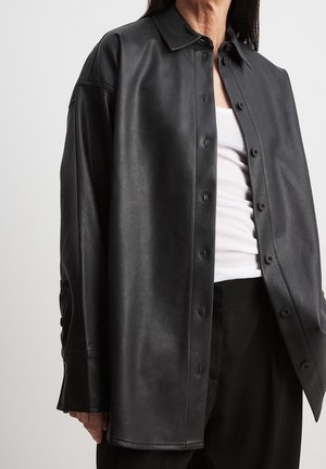 Person trägt ein übergroßes schwarzes Lederhemd mit Knöpfen über einem weißen Tanktop und schwarzen Hosen vor einem einfarbigen Hintergrund.