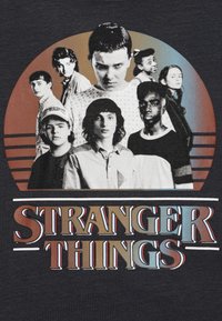 T-shirt en coton noir avec un motif imprimé représentant plusieurs personnages de "Stranger Things" avec un fond circulaire et du texte.