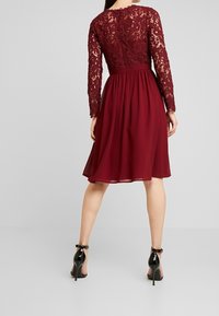 Bordové šaty s krajkovým korzetem a dlouhými rukávy, splývavá sukně z šifonu a vypasovaný pas. Nosí se s černými vysokými podpatky.