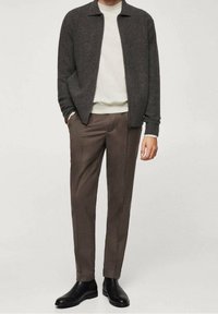 Giacca in maglia grigia scura indossata sopra un maglione a collo rotondo color crema, abbinata a pantaloni marroni su misura e stivali neri alla caviglia. Design semplice e moderno.