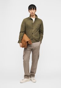 Άνδρας που φοράει χακί κουμπωτό σακάκι, μπεζ παντελόνια cargo, λευκά αθλητικά παπούτσια, κρατώντας διπλωμένο καφέ δερμάτινο σακάκι μπροστά σε λευκό φόντο.