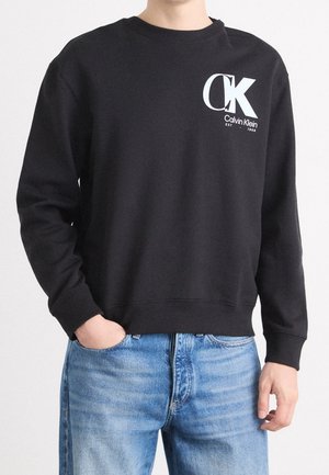 Osoba ubrana w czarną bluzę Calvin Klein z dużym białym logo "CK" na piersi, w połączeniu z jasnoniebieskimi dżinsami.