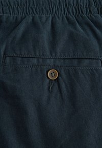 Un material de culoare albastru închis cu o textură netedă prezintă un brâu elastic și o singură buzunar la spate, asigurată cu un buton maro.