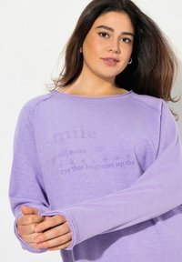 Maglietta a maniche lunghe di colore lilla con maniche raglan e una texture morbida, con testo che definisce "sorriso" stampato sul davanti.