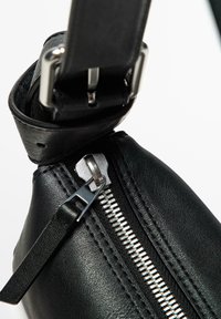Sac en cuir noir avec une fermeture éclair argentée, présentant une finition texturée, un tirette de fermeture éclair proéminente et un attache de sangle ajustable et large.