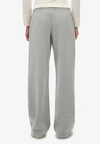 Persona vistiendo pantalones de chándal anchos en gris claro y una camiseta de manga larga en color crema, de pie con la espalda hacia la cámara contra un fondo blanco plano.