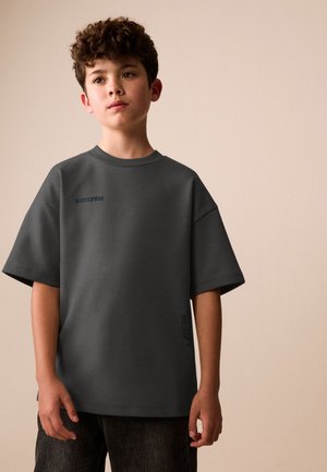 Garçon aux cheveux bouclés portant un t-shirt ample gris foncé et un pantalon noir, regardant légèrement vers le haut sur un fond beige uni.