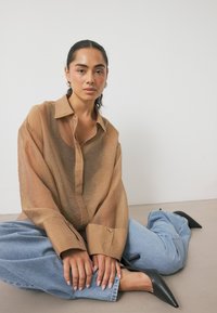 Svetlo rjava, polprosojna srajca s dolgimi rokavi in sproščenim krojem, v kombinaciji s svetlo modrimi širokimi džinsom in črnimi špicastimi petkami.