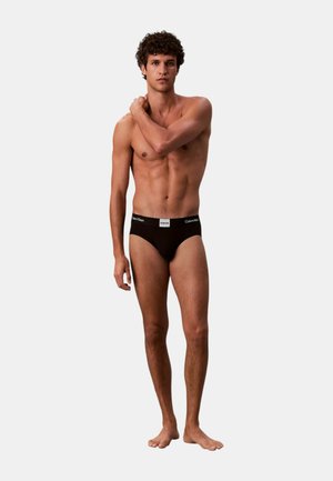 Slip nero in cotone con fascia in vita bianca con logo "Calvin Klein". Il modello è scalzo con una mano sulla spalla, su sfondo grigio.