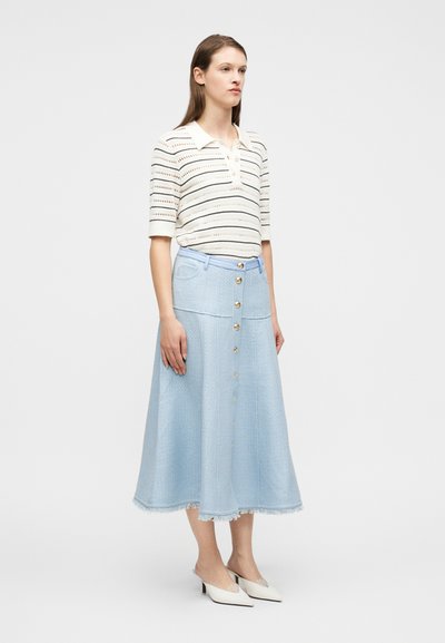 Γυναίκα φορώντας ριγέ πλεκτή κοντομάνικη μπλούζα, ανοιχτό μπλε midi φούστα με κουμπιά και τελείωμα με φουντίτσες, και λευκά μυτερά mules με χαμηλό τακούνι, στέκεται πλάγια.