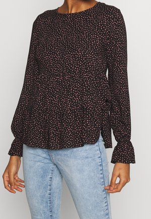 Vrouw draagt een zwarte blouse met lange mouwen met kleine roze stippen en lichtblauwe jeans, staand tegen een effen achtergrond.