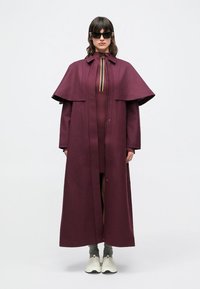 Burgundy long coat with oversized cape-style sleeves and a zip closure; paired with a fitted dress and light sneakers. Smooth texture.